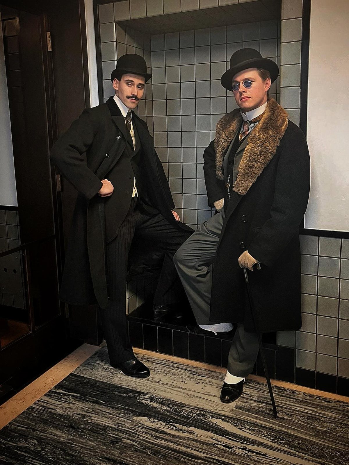 Two men dressed in vintage 1920s gangster costumes posing against a tiled wall.