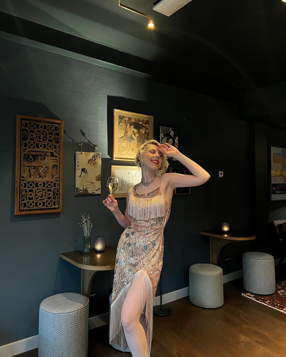 A blonde woman posing in a sequined 1920s-style evening dress with long fringe in a dark room.