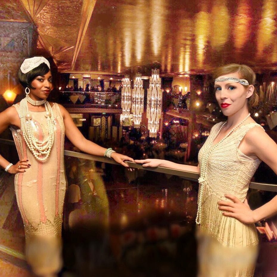 Two women wearing beaded flapper dresses and pearl necklaces posing in a golden bar.