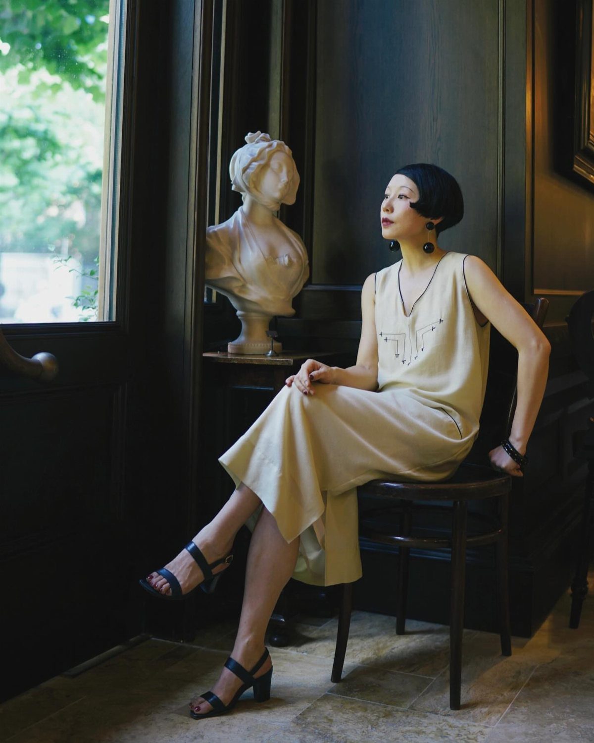 Woman in a cream drop-waist dress sitting beside a white bust sculpture in a dark room.