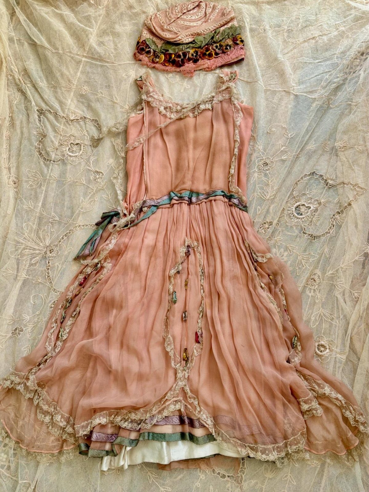 Peach vintage dress with lace trim and an embroidered hat laid on cream lace fabric.
