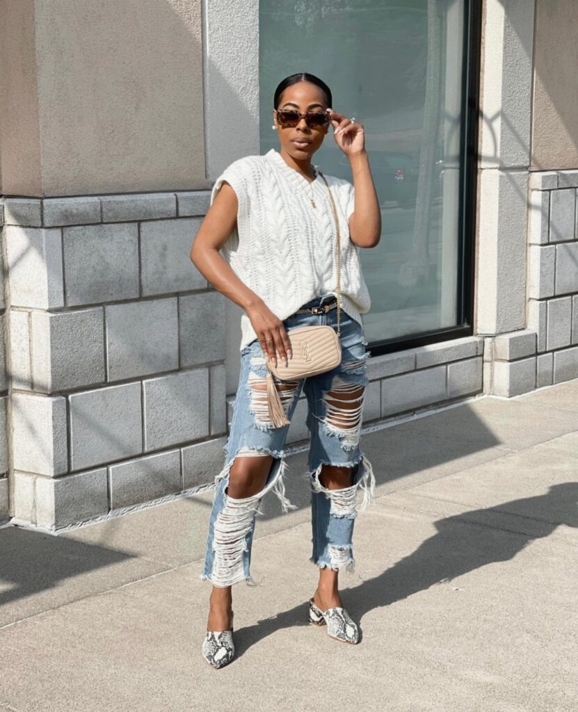 A woman posing in a white knit sweater and heavily distressed blue ripped jeans.