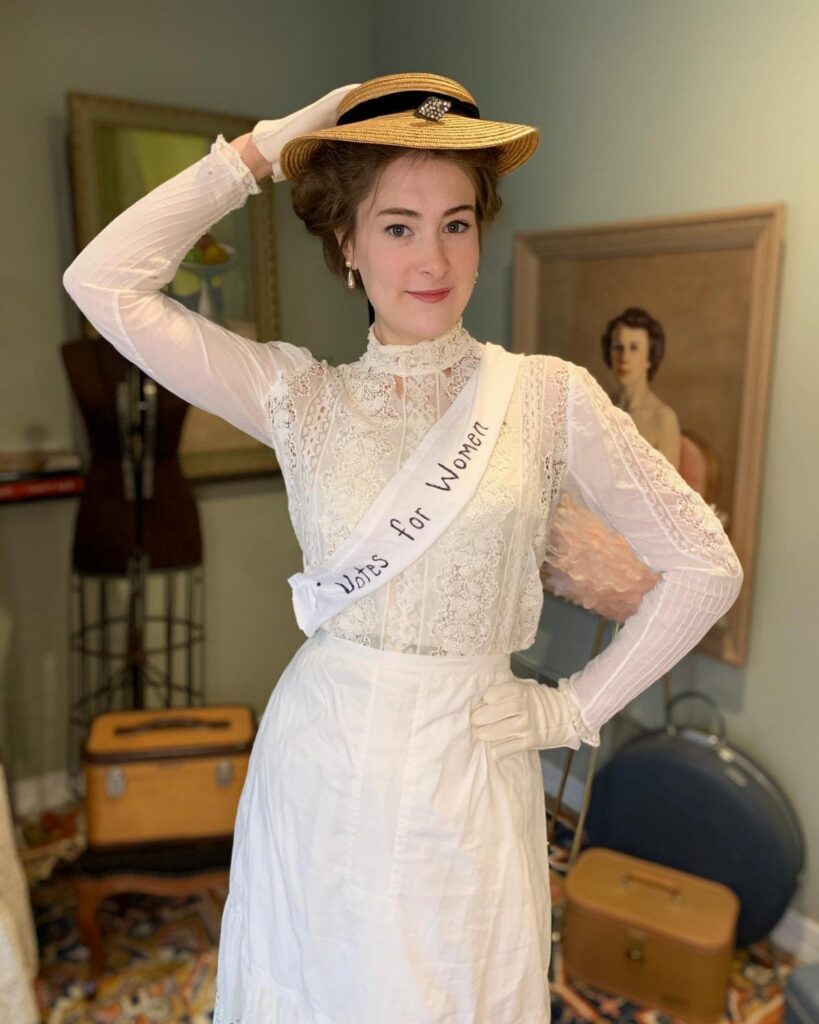 A woman in a white lace outfit and straw hat wearing a Votes for Women sash.