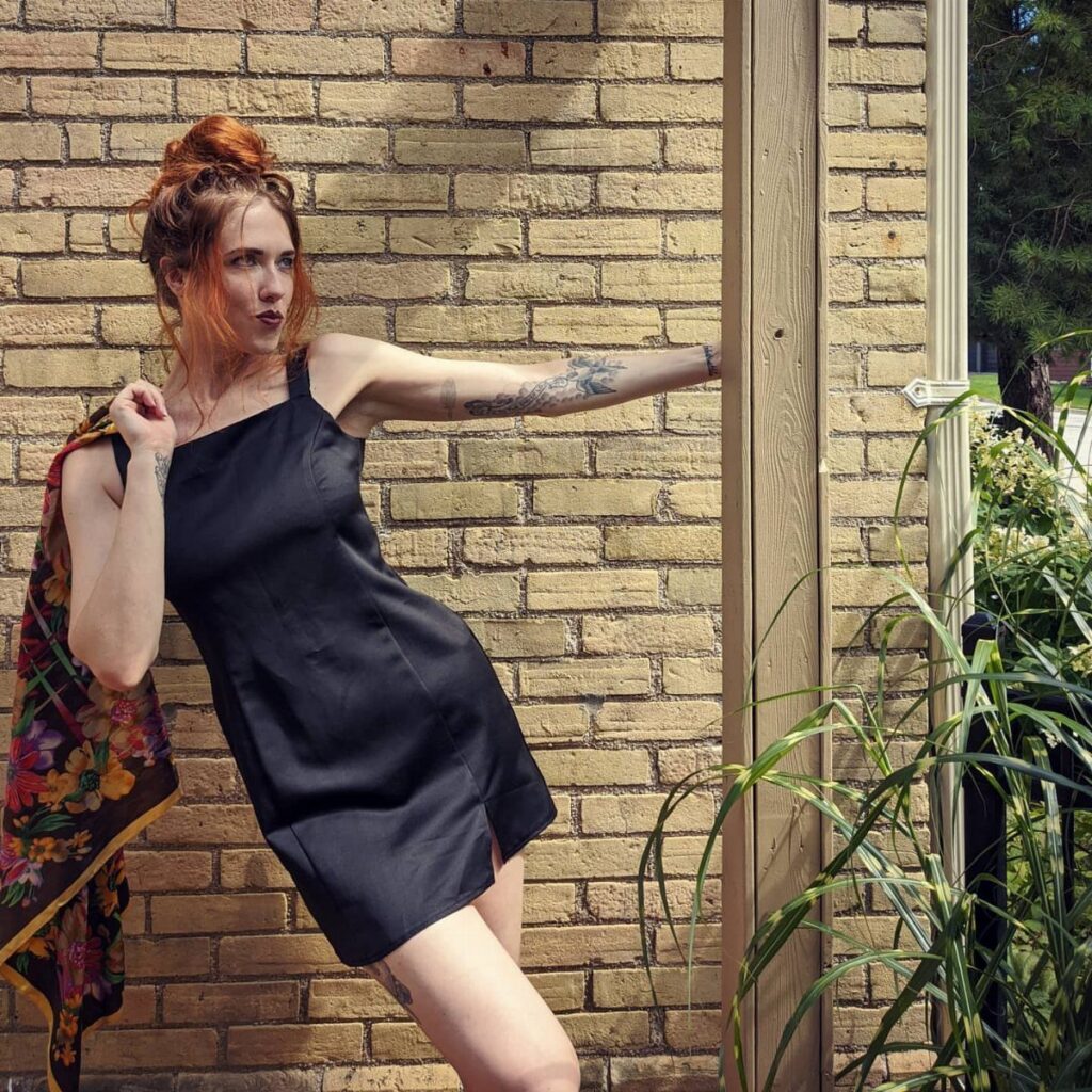 Woman with red hair wearing a black satin slip dress against a brick wall.