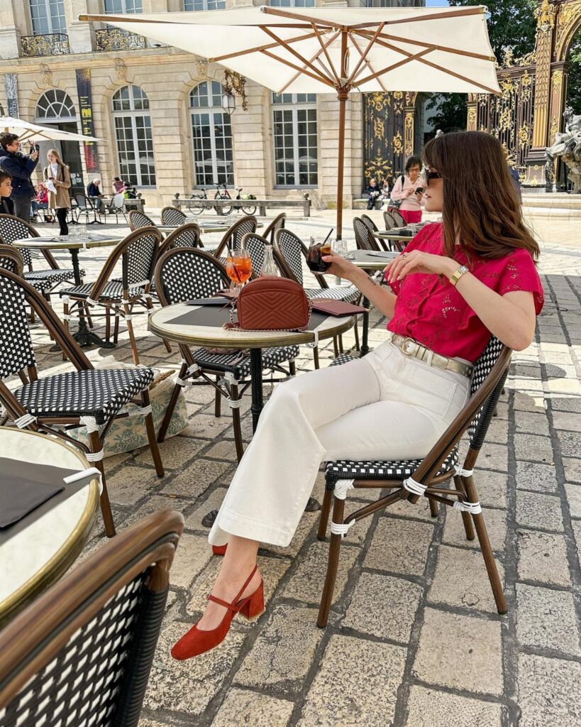 Woman in a pink blouse and white trousers sitting at an outdoor cafe table.