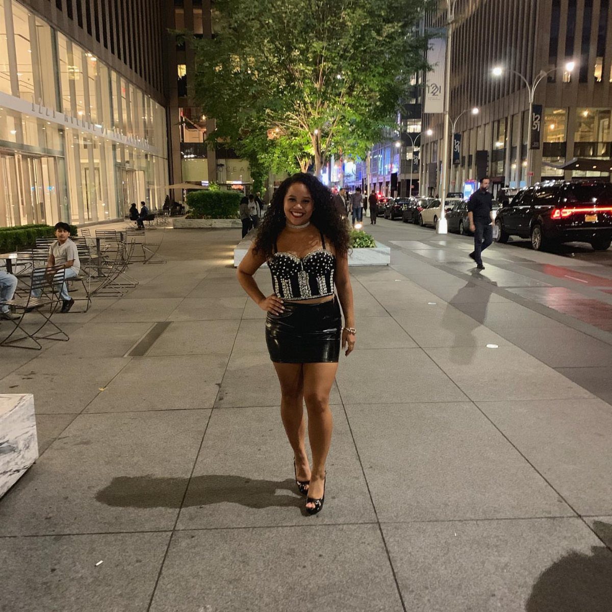 A woman posing in a sequined crop top and black leather mini skirt on a city sidewalk.