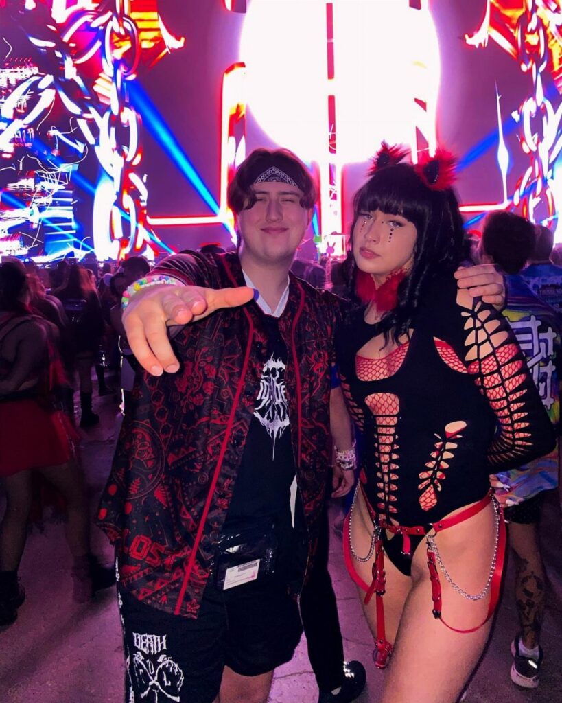 A young couple posing in black and red rave outfits at a neon-lit music festival.