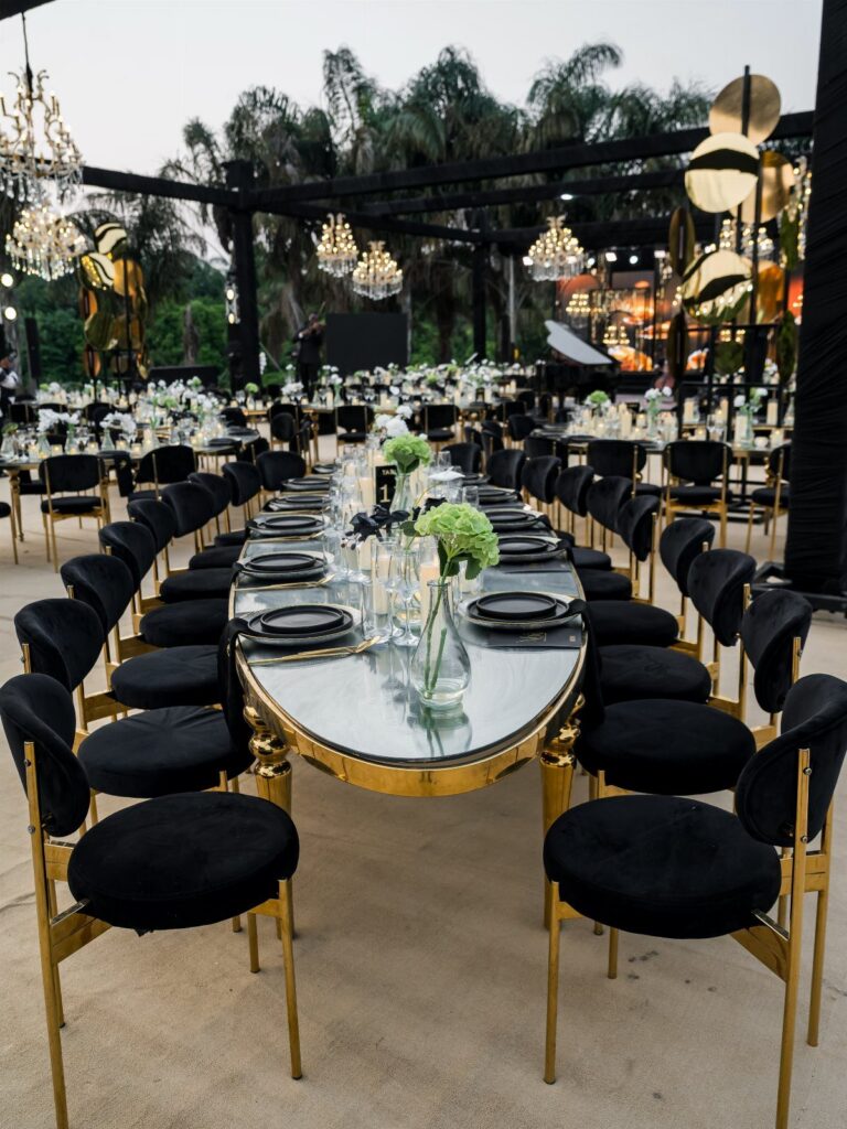 Black velvet and gold chairs arranged around long glass-topped banquet tables at an outdoor event.
