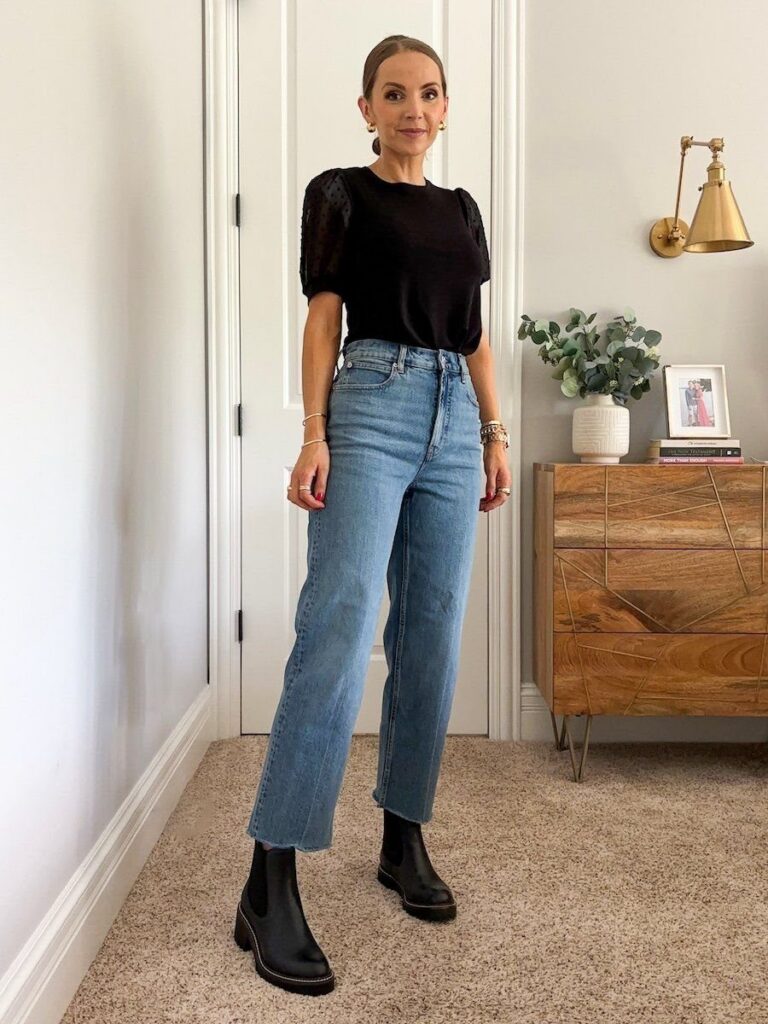Woman wearing a black top, blue jeans, and black leather Chelsea boots standing indoors.