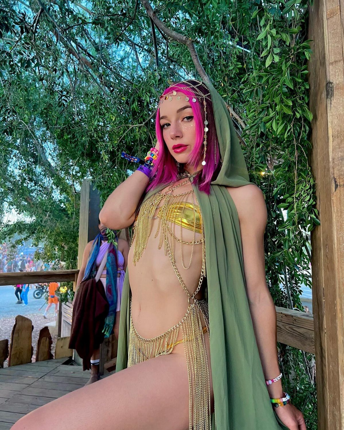Woman with pink hair wearing a gold fringed bodysuit and green hooded cape outdoors.