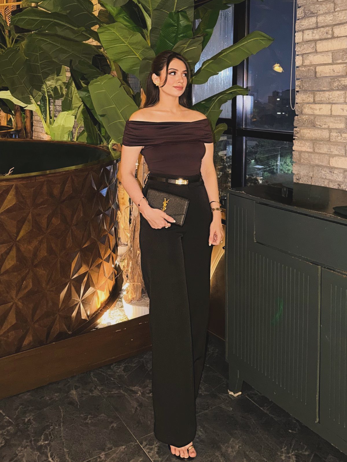 A woman posing in a dark brown off-the-shoulder top and black wide-leg trousers.