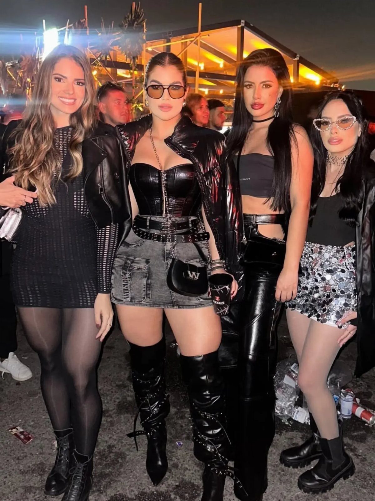 Four women posing in black leather and denim festival outfits at an outdoor night event.