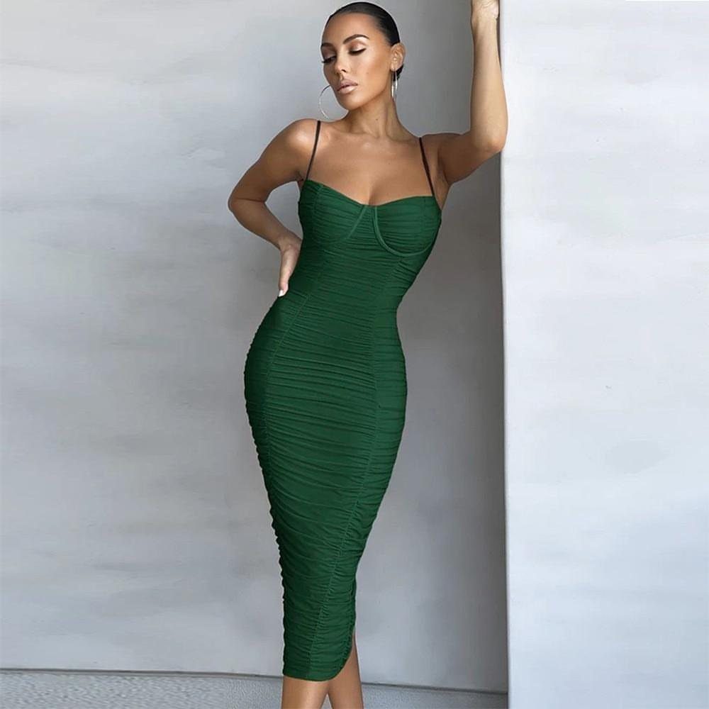 A woman in a tight emerald green ruched dress posing against a white wall.