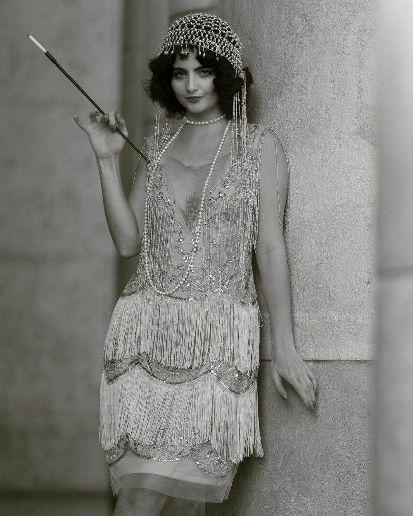 A woman wearing a beaded flapper costume and headband holding a long cigarette holder.