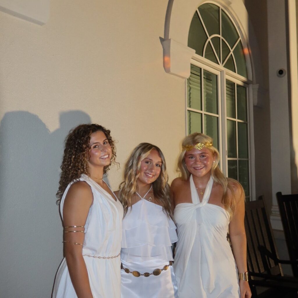 Three young women posing in white toga-style dresses and gold accessories at night.
