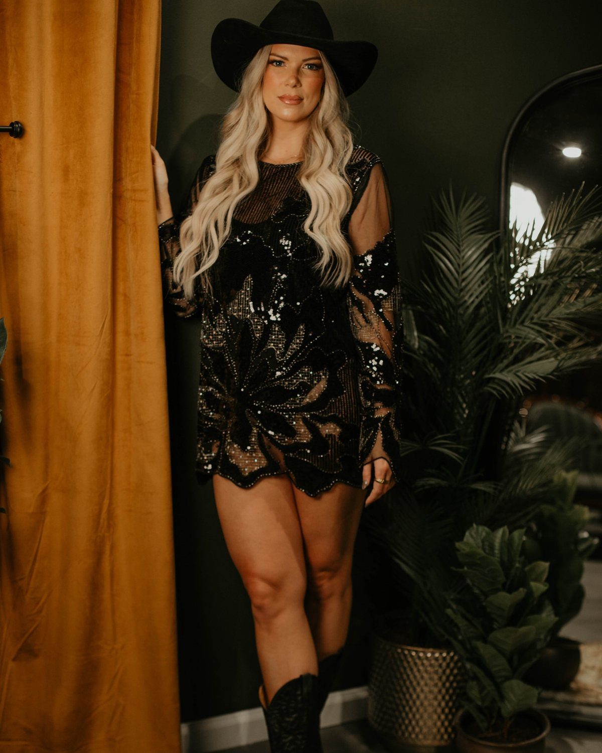 A blonde woman posing in a black sequined mini dress, cowboy hat, and black boots.