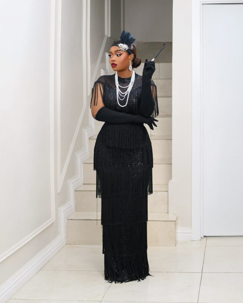 A woman in a black fringed flapper dress, long gloves, and pearls stands by a staircase.