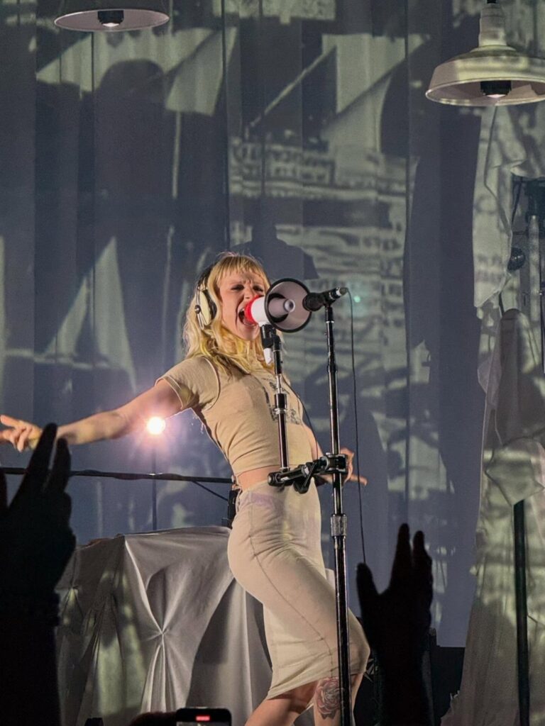 A blonde singer in a tan outfit performs on stage with a microphone and headphones.