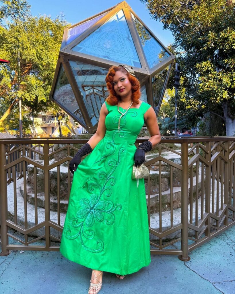 A woman in a green embroidered gown and black gloves posing outdoors near a glass sculpture.