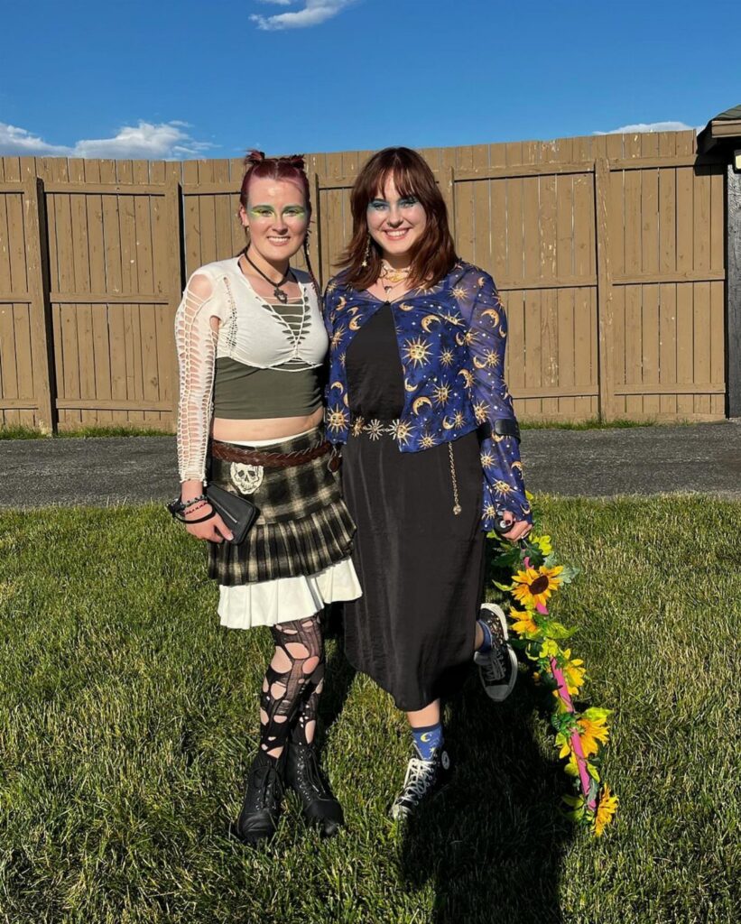 Two women posing in bohemian style outfits in a grassy yard by a wooden fence.