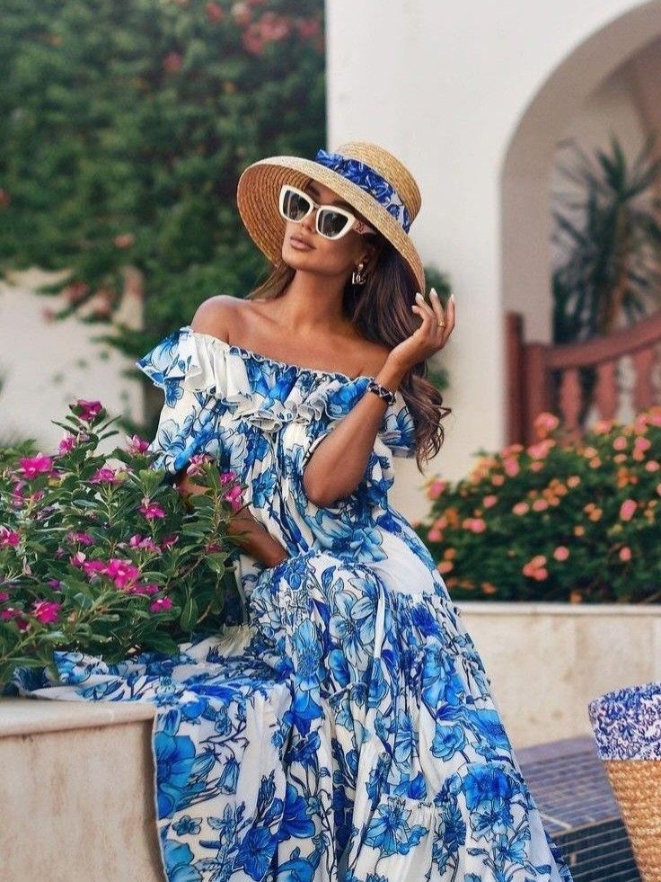 Woman in a blue floral maxi dress and straw hat sitting among garden flowers.