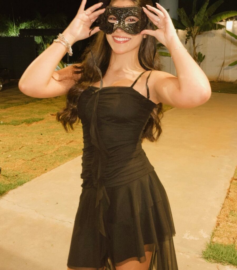 A smiling woman wearing a black sequined mask and a black dress posing outdoors at night.