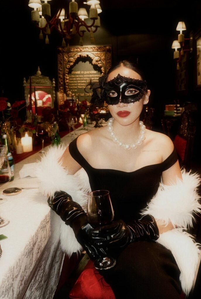 Woman in a black masquerade mask, black dress, and white fur sitting at a banquet table.