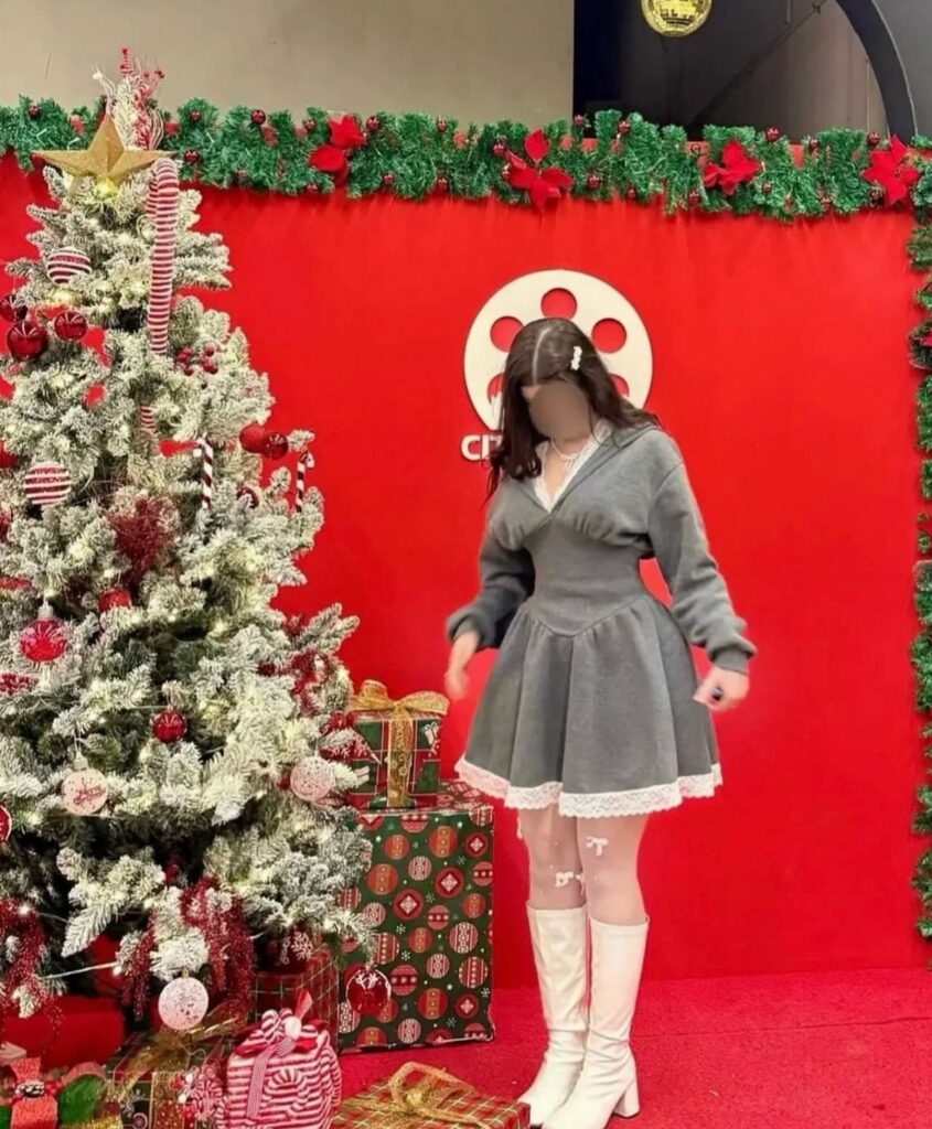 A person wearing a grey mini dress, white tights, and white knee-high boots by a Christmas tree.