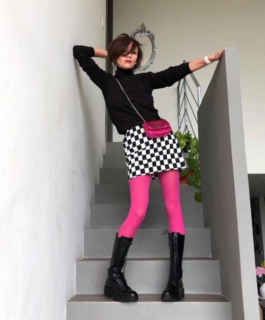 A person posing on stairs wearing a checkered skirt and bright pink leggings with black boots.
