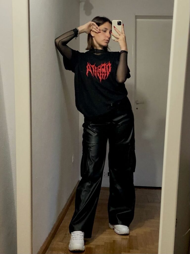 Woman in black graphic t-shirt and faux leather trousers posing for a mirror selfie.