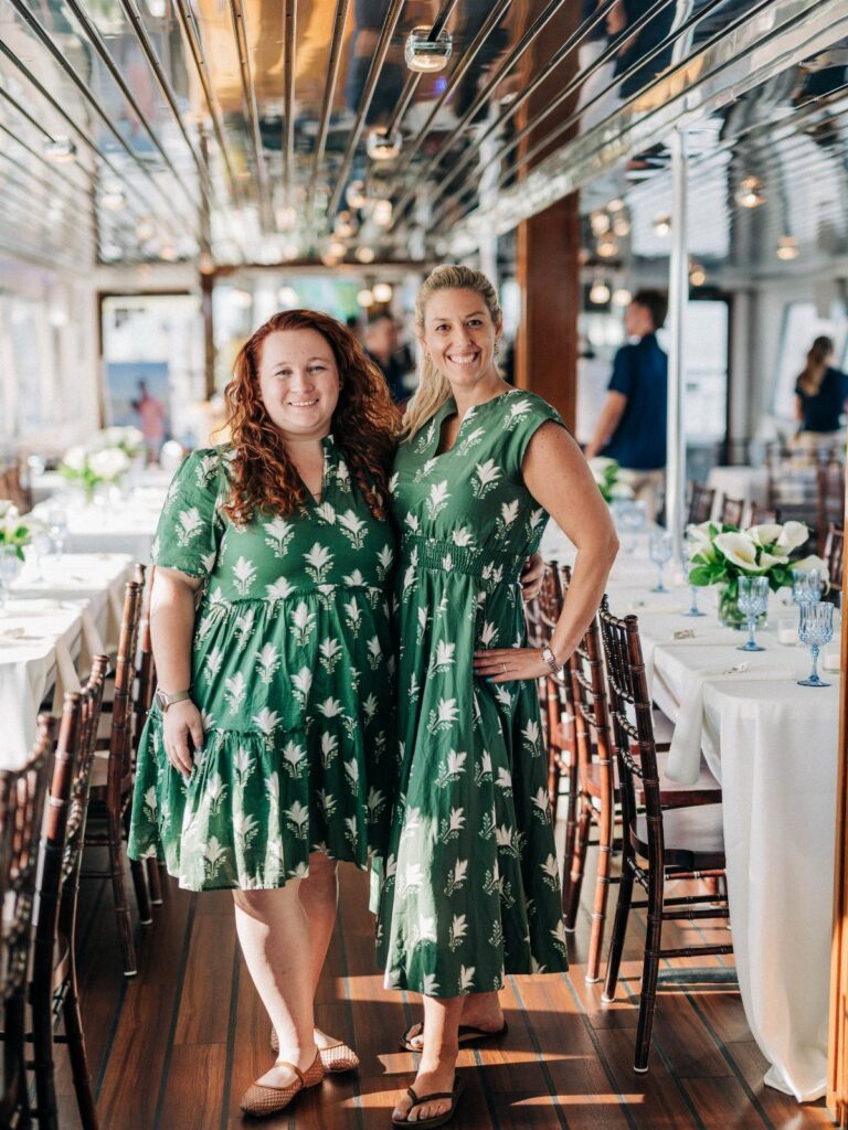 Two women in green patterned dresses posing together in a decorated dining room.