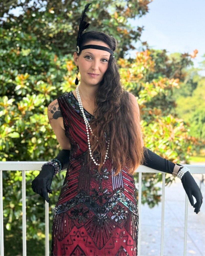 A woman in a sequined flapper dress and pearl necklace posing by a white railing.