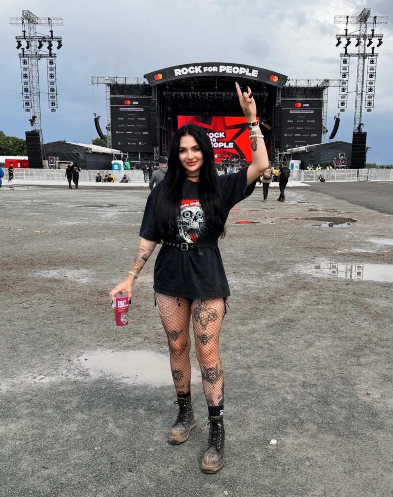A woman in a black graphic t-shirt, fishnet tights, and combat boots posing at a music festival.