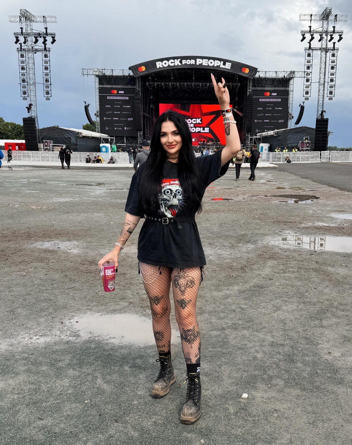 A woman in a black graphic t-shirt, fishnet tights, and combat boots posing at a music festival.