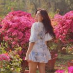 A woman in a white floral sundress standing amidst bright pink bougainvillea flowers in a garden.
