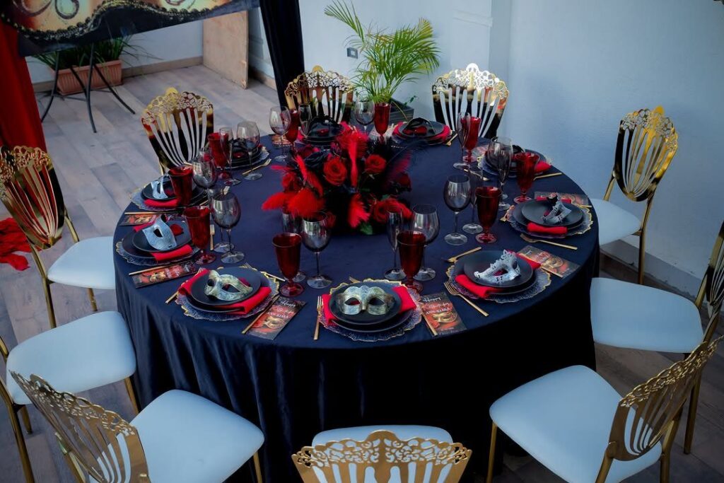 A formal dinner table set with black linens, red roses, feathers, and masquerade masks.