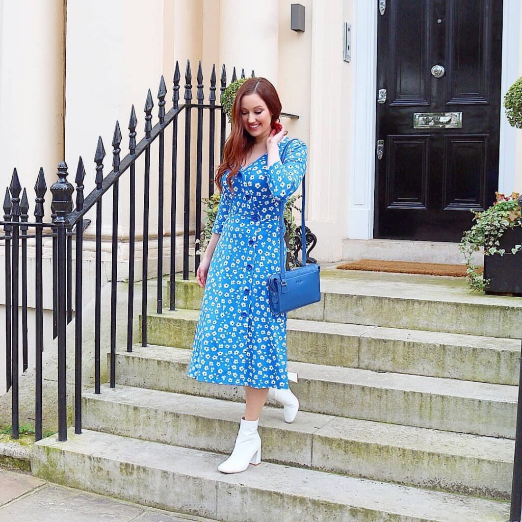 Woman in a blue floral midi dress and white boots standing on outdoor stone steps.