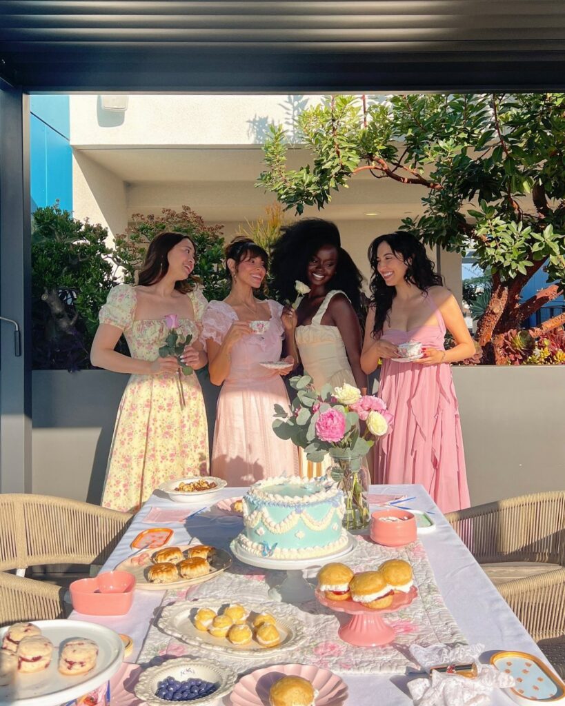 Four women in floral and pastel dresses standing behind a decorated tea party table outdoors.
