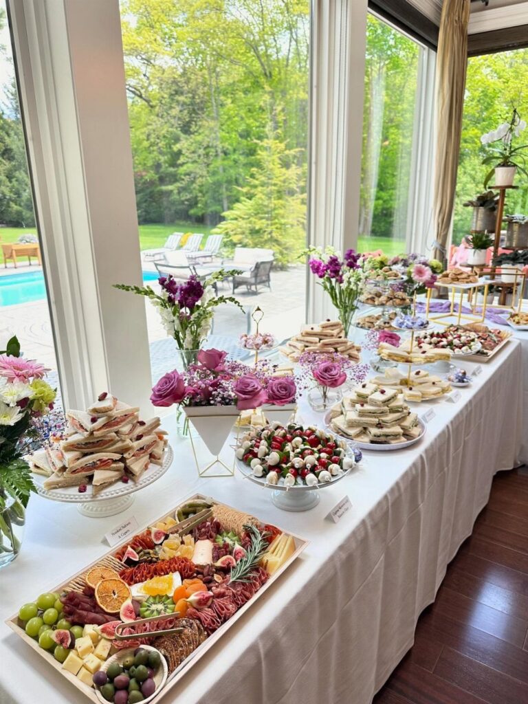 A long buffet table filled with tea party finger foods and floral arrangements near a large window.