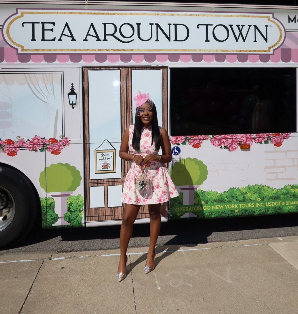 A woman in a pink floral dress and fascinator posing in front of a tea food truck.