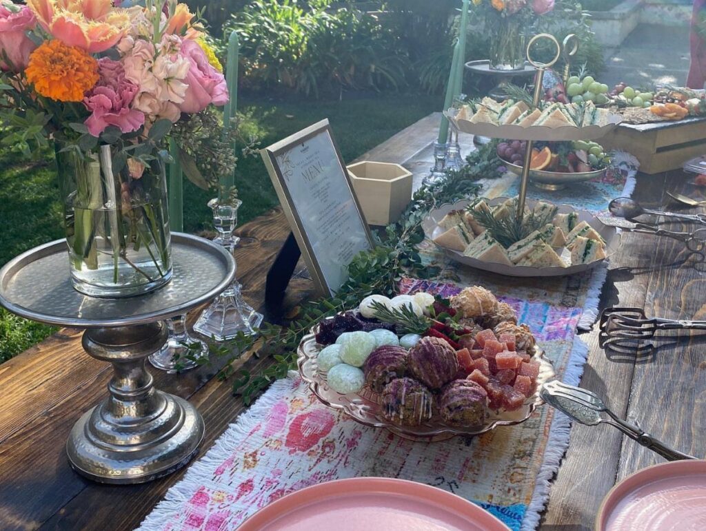 A long wooden table set with floral arrangements, tiered sandwich trays, and savory appetizers in a sunny garden.