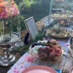 A long wooden table set with floral arrangements, tiered sandwich trays, and savory appetizers in a sunny garden.