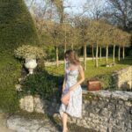 A woman wearing a white floral dress sits on a stone wall in a garden.