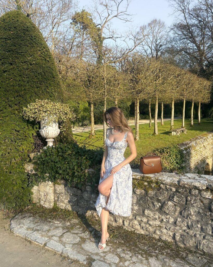 A woman wearing a white floral dress sits on a stone wall in a garden.