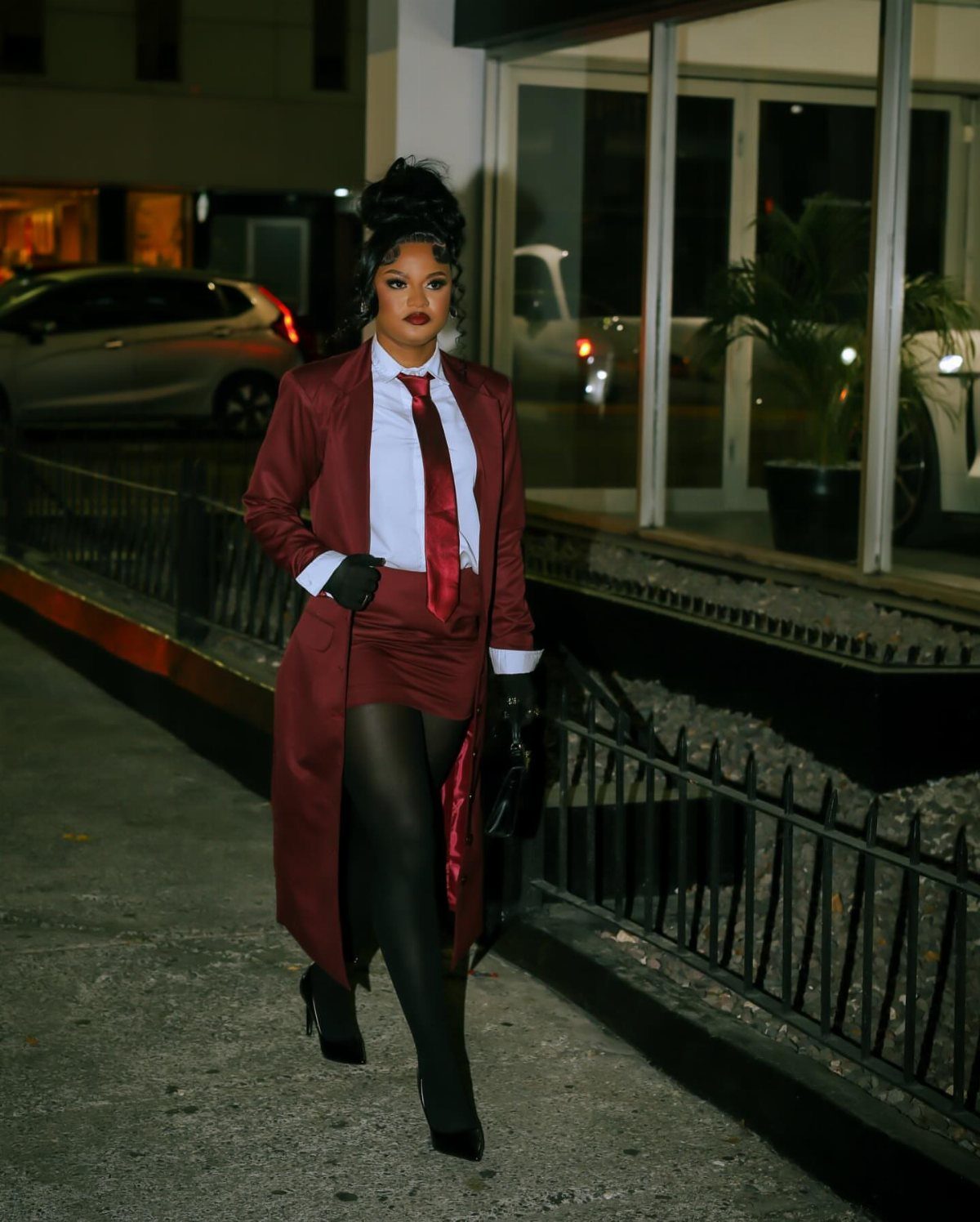 A woman in a maroon suit, long coat, and red tie posing on a city sidewalk at night.