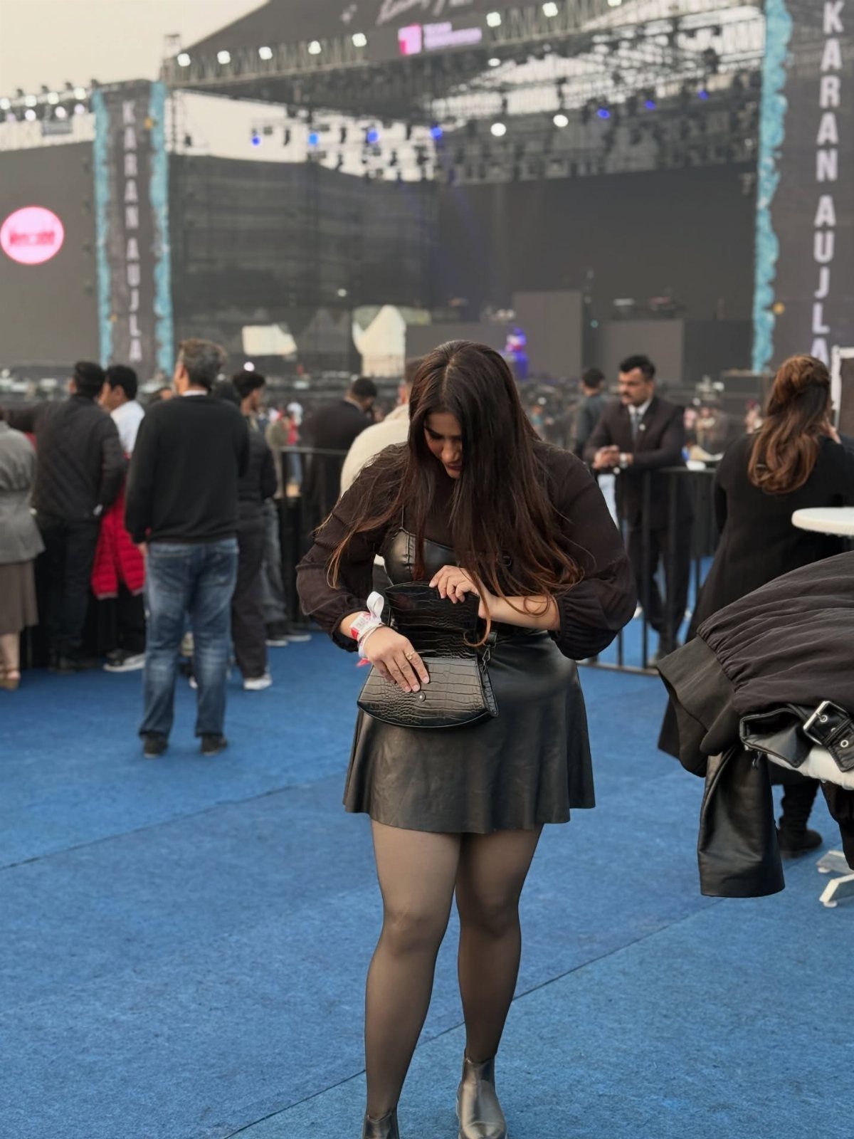 A woman in a black leather skirt, black tights, and silver boots stands at an outdoor concert.
