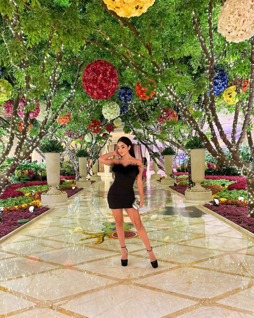 A woman in a black dress posing in a luxurious indoor garden with colorful floral decorations.
